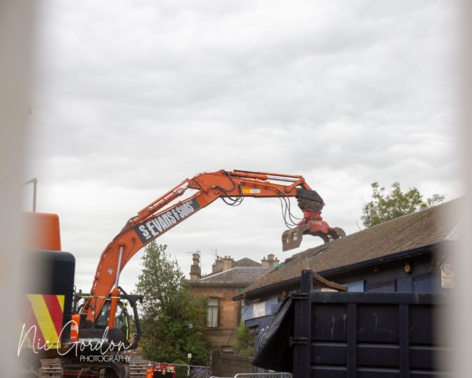 A large excavator grabbing the roof of the old booking hall